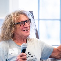 Joe Paterick smiles as he hold microphone in right hand and raises his left while talking